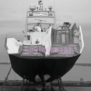 May include: A black and white photograph of a large boat on land. The boat is on blocks and has a large propeller. The boat has a white top and a black bottom.