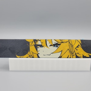 Può includere: Un braccialetto in pelle nera con un piccolo foro sul lato sinistro presenta un design stampato di un personaggio in stile anime con lunghi capelli biondi e occhi blu in nero e giallo.