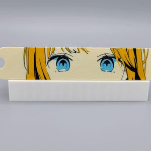 May include: A close-up of a white rectangular bookmark with a colorful anime-style illustration of a woman's eyes. The bookmark has a hole in the top left corner.