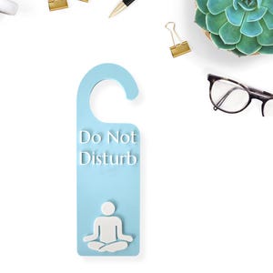May include: A light blue door hanger with the words "Do Not Disturb" in white. Below the text is a white graphic of a person in a seated yoga pose. The hanger is surrounded by office supplies and a succulent.