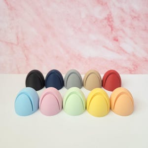 May include: A collection of ten colorful egg-shaped place card holders. The holders are in various colors, including black, navy, gray, tan, red, light blue, pink, green, yellow, and orange. Each holder has a slot at the top for a card.