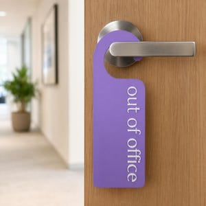 May include: A purple door hanger with the words "out of office" in white text. The door hanger is hanging on a wooden door with a silver handle. The background shows a hallway with a plant.