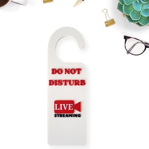 May include: A white door hanger with a curved top and the words "DO NOT DISTURB" in red. Below, a red rectangle with "LIVE" and a video camera icon, and "STREAMING" in black. Ideal for a content creator.