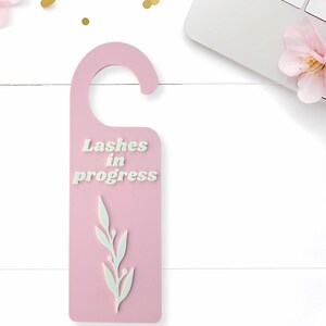May include: A pink door hanger with the text "Lashes in progress" in white. A white leaf design is below the text. The door hanger is near a laptop and pink flowers.