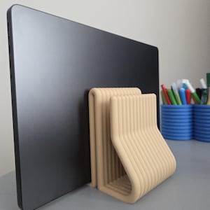 May include: A beige, ribbed, U-shaped laptop stand holding a thin, dark gray laptop in an upright position. The stand features a series of parallel, vertical ridges along its exterior surface, providing a stable base for the electronic device.