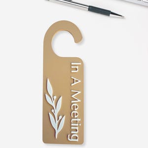 May include: A gold-colored door hanger with the text "In A Meeting" in white. A white leaf design is on the left side. A pen and keyboard are in the background.