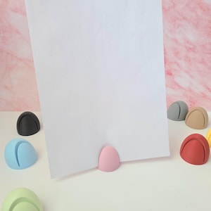 May include: A white sheet of paper held by a pink egg-shaped holder. Several colorful, egg-shaped holders in black, blue, green, gray, tan, red, and yellow are scattered around. The background is a pink marble pattern.