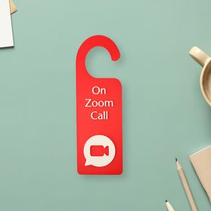 May include: A red door hanger with the text "On Zoom Call" and a white speech bubble with a video camera icon. The door hanger is designed to be hung on a door handle.