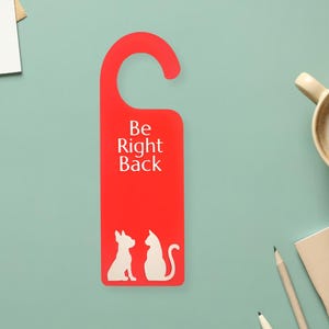 May include: A red door hanger with the text "Be Right Back" in white. Below the text are white silhouettes of a dog and a cat. The hanger is designed to be hung on a door handle.