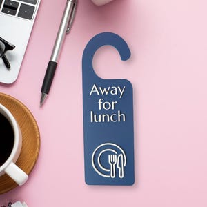 May include: A navy blue door hanger with the text "Away for lunch" in white. The bottom of the hanger features a white plate and silverware graphic. The hanger is on a pink surface with a pen, a laptop, and a cup of coffee.