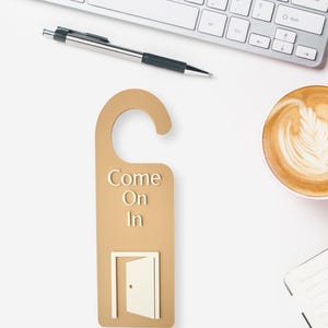 May include: A tan door hanger with the words "Come On In" and a graphic of an open door. A silver pen, a keyboard, and a cup of coffee are also visible on a white surface.