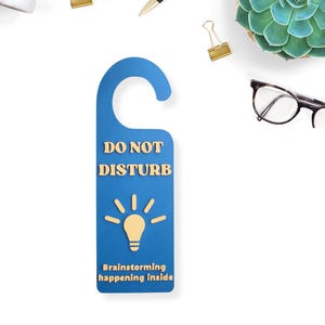 May include: A blue door hanger with the words "DO NOT DISTURB" in gold lettering. Below the text is a lightbulb graphic and the words "Brainstorming happening inside". The hanger is surrounded by office supplies and a succulent.