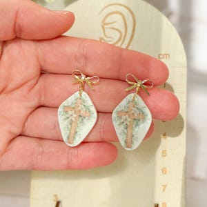 May include: Earrings with a cross design. Each earring has a gold bow and a diamond-shaped pendant with a cross and floral pattern. The background is a light green colour. The earrings are approximately 2 cm in length.