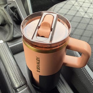 Könnte beinhalten: Ein beigefarbener Stanley-Reisebecher mit schwarzem Boden und braunem Griff, platziert in einem Getränkehalter im Auto. Der Deckel ist hellbraun mit einem Klappverschluss. Das Stanley-Logo ist sichtbar.