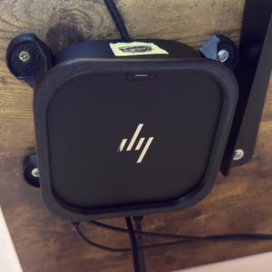 May include: A black square electronic device with a silver HP logo on the front, mounted to a wood-grain surface using a black plastic bracket. The device features a small indicator light at the top and black cables extending from the bottom.