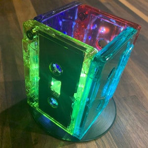 Peut inclure: Une pile de quatre cassettes audio transparentes, chacune d'une couleur diff&eacute;rente&nbsp;: vert, bleu, rouge et violet. Les cassettes sont empil&eacute;es les unes sur les autres, la cassette verte en bas et la cassette violette en haut.