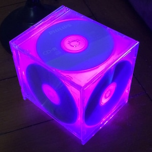 May include: A clear plastic cube with four CD-Rs inside. The cube is lit from the inside with a purple light, making the CDs glow.
