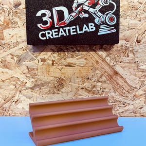 Könnte beinhalten: Ein terrakottafarbenes 3D-gedrucktes Objekt mit geschwungenem Design, das auf einer hellblauen Oberfläche ruht. Darüber befindet sich ein schwarzes Schild mit dem Text "3D CREATELAB" und einer Grafik eines Roboterarms.