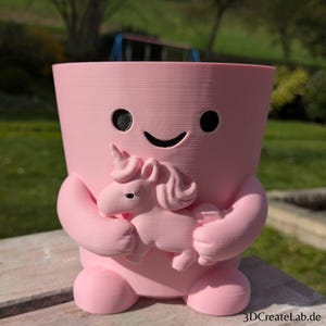 Könnte beinhalten: Ein rosa Blumentopf mit einem lächelnden Gesicht und einem kleinen Einhorn-Design. Der Topf hat Arme und Beine und hält ein kleines Einhorn. Der Topf besteht aus einem strukturierten Material.