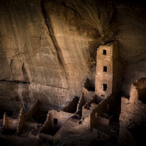 May include: An ancient stone structure built into a cliff face. The structure includes a tall tower with three visible windows and several lower, connected rooms. The scene is lit by natural light, creating shadows and highlighting the texture of the stone.