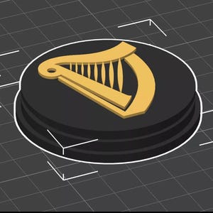 Op de afbeelding: Een zwart en goud rond embleem met een gouden harp ontwerp. Het embleem is verhoogd op een zwarte cirkelvormige basis met een witte rand. De achtergrond is een donkergrijze raster.