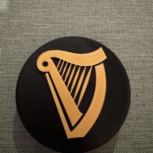 Op de afbeelding: Een zwarte, ronde onderzetter met een gouden harp ontwerp. De harp is een gestileerde weergave van het instrument. De onderzetter is waarschijnlijk gemaakt van een duurzaam materiaal, om oppervlakken te beschermen tegen drank.