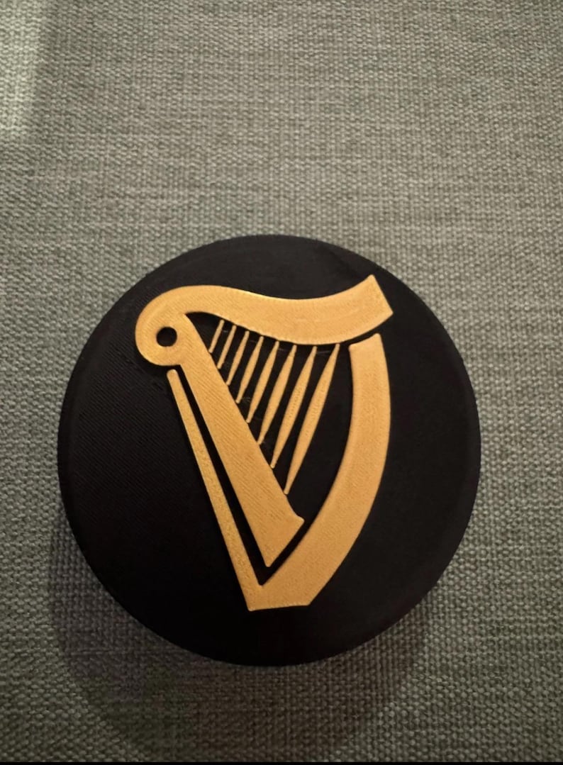 Op de afbeelding: Een zwarte, ronde onderzetter met een gouden harp ontwerp. De harp is een gestileerde weergave van het instrument. De onderzetter is waarschijnlijk gemaakt van een duurzaam materiaal, om oppervlakken te beschermen tegen drank.