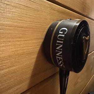 Op de afbeelding: Een zwarte Guinness biertap met gouden accenten en het Guinness-logo, gemonteerd op een houten muur. De tap heeft een lange tuit en een harpsymbool.