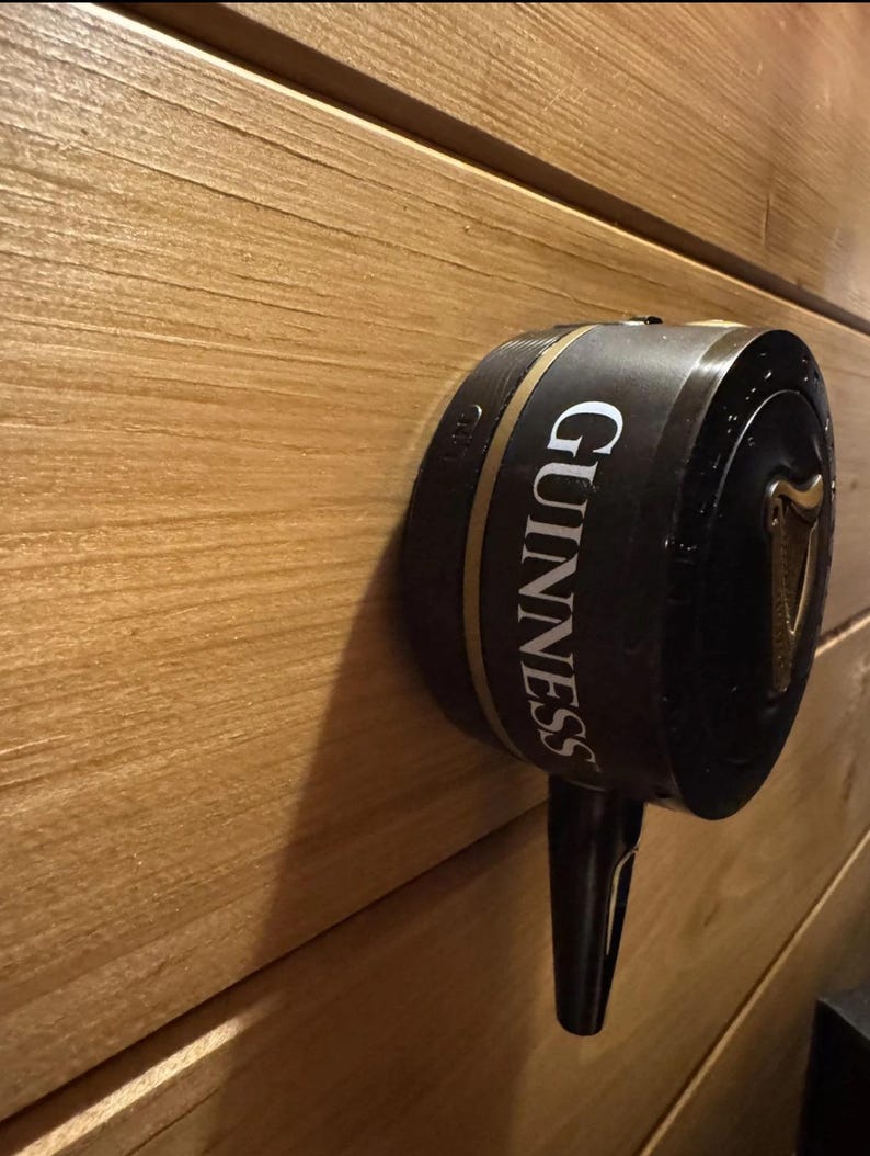 Op de afbeelding: Een zwarte Guinness biertap met gouden accenten en het Guinness-logo, gemonteerd op een houten muur. De tap heeft een lange tuit en een harpsymbool.