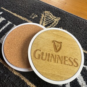May include: Two round coasters. One has a cork base, the other has a light wood surface with the Guinness logo and text. Both have a white border. The coasters are on a dark gray patterned surface.