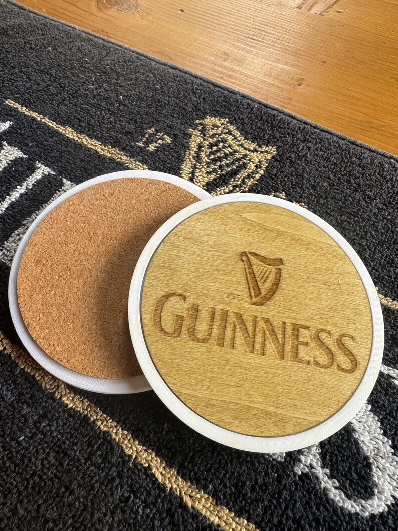 May include: Two round coasters. One has a cork base, the other has a light wood surface with the Guinness logo and text. Both have a white border. The coasters are on a dark gray patterned surface.