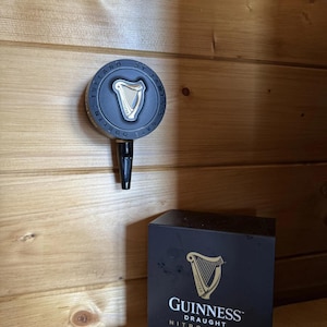 Op de afbeelding: Een zwarte en zilveren Guinness tapkraan met een harp logo, gemonteerd op een houten muur. Daaronder staat een zwarte doos met het Guinness logo en de tekst "NitroSurge".