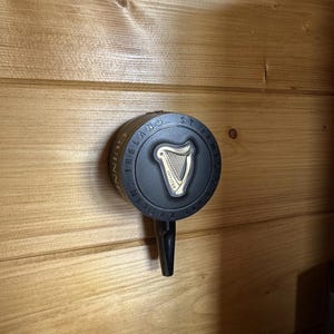 Op de afbeelding: Een zwarte Guinness biertap met een cirkelvormige voorkant met het Guinness-logo, een gouden harp en de woorden "Guinness Ireland St. James's Gate Dublin 1759". De tap is op een houten muur gemonteerd.