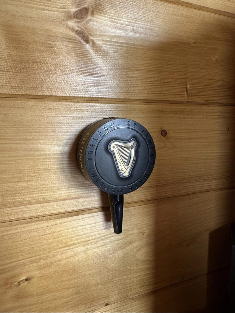 Op de afbeelding: Een zwarte Guinness biertap met een cirkelvormige voorkant met het Guinness-logo, een gouden harp en de woorden "Guinness Ireland St. James's Gate Dublin 1759". De tap is op een houten muur gemonteerd.