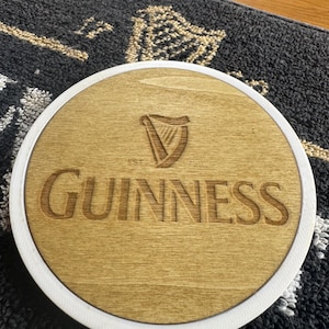May include: A round wooden coaster with a white border. The coaster features the Guinness logo, a harp, and the brand name in a carved, light brown finish. The coaster is on a patterned surface.