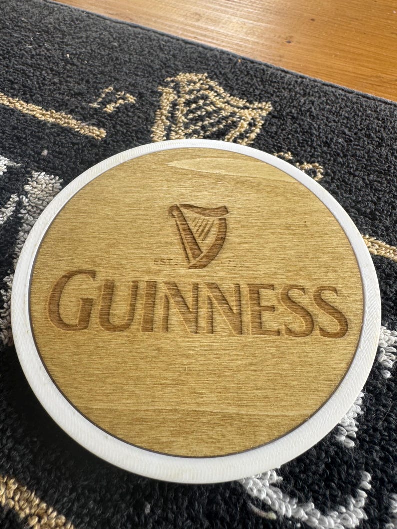 May include: A round wooden coaster with a white border. The coaster features the Guinness logo, a harp, and the brand name in a carved, light brown finish. The coaster is on a patterned surface.