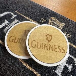 May include: Two round wooden coasters with the Guinness logo, featuring the brand name and a harp symbol, are displayed. The coasters have a light wood center with a white border. They rest on a dark gray mat.