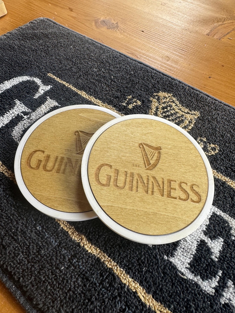 May include: Two round wooden coasters with the Guinness logo, featuring the brand name and a harp symbol, are displayed. The coasters have a light wood center with a white border. They rest on a dark gray mat.
