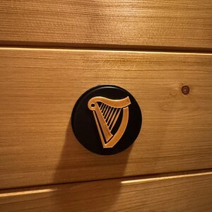 Op de afbeelding: Een zwart, rond embleem met een gouden harpontwerp, bevestigd op een lichtbruin houten oppervlak. De harp is een traditioneel Iers symbool. Het hout heeft een horizontale nerf.