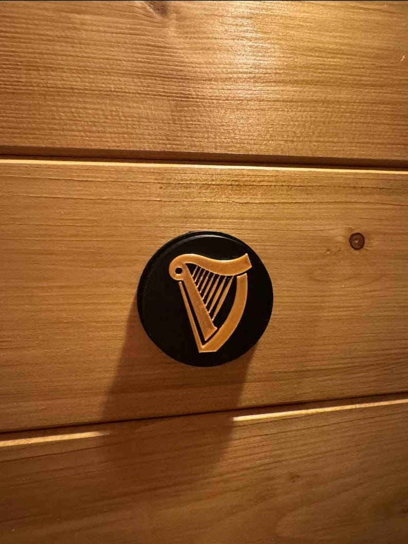 Op de afbeelding: Een zwart, rond embleem met een gouden harpontwerp, bevestigd op een lichtbruin houten oppervlak. De harp is een traditioneel Iers symbool. Het hout heeft een horizontale nerf.
