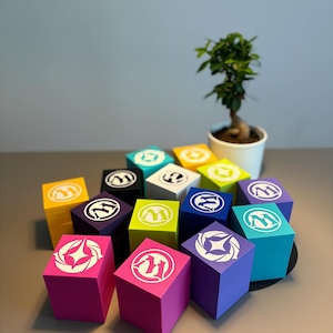 Op de afbeelding: Een verzameling kleurrijke, vierkante dozen met witte logo's. De dozen zijn in verschillende tinten, waaronder roze, geel, blauw en groen. Een kleine bonsai-boom in een witte pot staat op de achtergrond. De dozen zijn op een grijze ondergrond geplaatst.