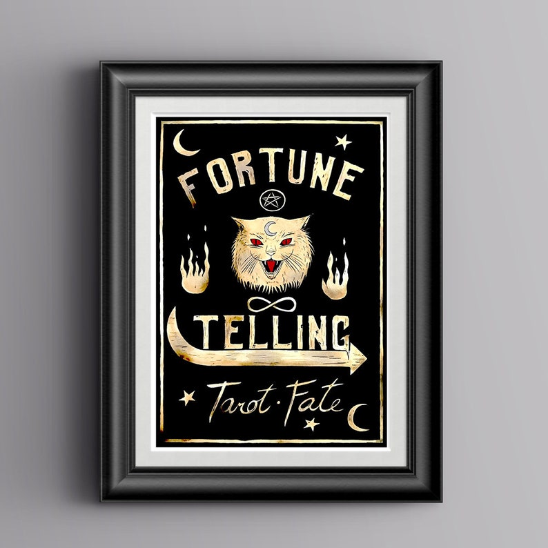 May include: A black and gold framed print with a black background and gold text that reads "Fortune Telling Tarot. Fate." A white cat with red eyes and a crescent moon on its forehead is in the center of the print. The cat is surrounded by flames and an infinity symbol.