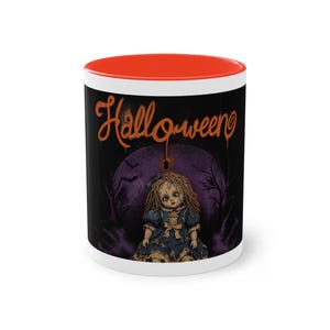 Könnte beinhalten: Ein Keramikbecher mit leuchtend orangefarbenem Innenraum und Rand. Die Außenseite zeigt einen schwarzen Hintergrund mit dem orangefarbenen Schriftzug Halloween über einer Grafik einer Puppe im blauen Kleid, einem violetten Mond, kahlen Bäumen.