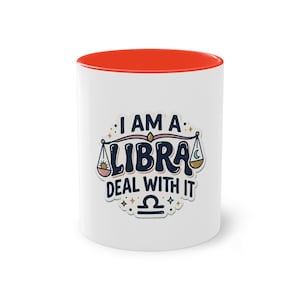 Könnte beinhalten: Ein weißer Keramikbecher mit leuchtend orangefarbenem Innenraum. Die Vorderseite zeigt eine Grafik mit dem Text I AM A LIBRA DEAL WITH IT in Dunkelblau, ergänzt durch eine Waagen-Illustration, kleine Sterne und das Waage-Tierkreiszeichen.