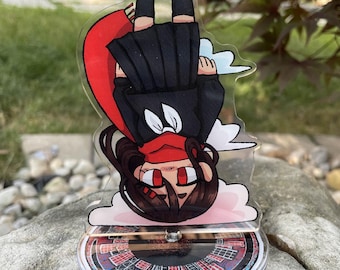 Puede incluir: Un expositor acrílico transparente con un personaje de dibujos animados con uniforme negro y sombrero rojo, con cabello castaño y ojos rojos. El expositor está sobre una base circular con un diseño de ciudad colorido.