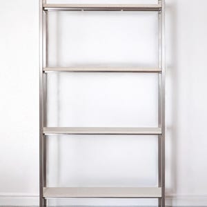 Könnte beinhalten: Ein modernes Bücherregal mit vier Regalböden, silberfarbenem Metallrahmen und hellen Regalböden. Die Regalböden sind gleichmäßig angeordnet, und das Bücherregal steht vor einer weißen Wand. Das Design ist minimalistisch und funktional.