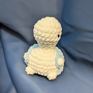 May include: White crocheted stuffed animal with blue accents. The animal is sitting on a blue fabric surface.