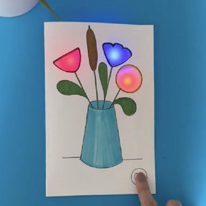 Puede incluir: Una tarjeta blanca con un dibujo de un jarrón azul que contiene un ramo de flores. Las flores son rojas, azules y naranjas y tienen bombillas en su interior. Hay un pequeño botón redondo en la esquina inferior derecha de la tarjeta.