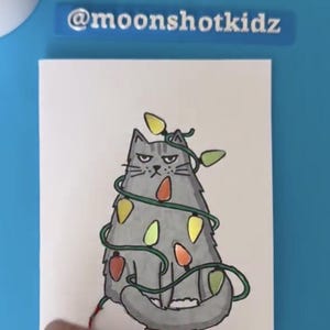 May include: A hand-drawn illustration of a gray cat wrapped in Christmas lights. The cat has a grumpy expression. The image includes the text "@moonshotkidz" at the top and bottom.