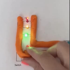 May include: A Play-Doh squishy circuit demonstration. Orange Play-Doh is molded into a circuit shape with LED lights. The lights are red, yellow, and green. The word "Switch" is printed on the paper. The text "@moonshotkidz" is visible.